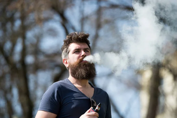 Concepto de cigarrillo electrónico. El hombre con barba larga y nubes de humo se ve relajado. El hombre barbudo fuma vapor en un día soleado. Hombre con barba y bigote en la cara tranquila, ramas en el fondo, desenfocado —  Fotos de Stock