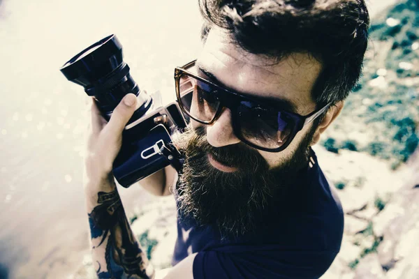 Funny tourist. Man with beard and mustache wears sunglasses, water surface on background. Guy shooting nature near river or pond. Hipster on smiling face holds old fashioned camera. Tourist