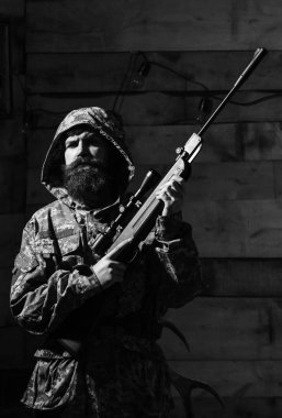 Cesur adam. Acımasız adam. Sakallı adam başlıklı kamuflaj elbiseler, ahşap iç arka plan giyer. Hunter, silahla acımasız hipster hazırlamak tüfek avcılık için