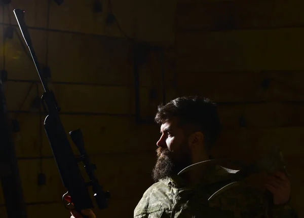 Hunter concept. Macho on strict face at gamekeepers house ready for hunting. Hunter brutal with gun and horns of deer, lighted in darkness. Man with beard wears camouflage clothing, dark background — Stock Photo, Image