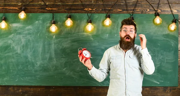 Esqueci-me do conceito do tempo. Homem com barba e bigode no rosto chocado na sala de aula. O professor de óculos tem despertador. Barbudo hipster detém relógio, quadro no fundo, espaço de cópia — Fotografia de Stock