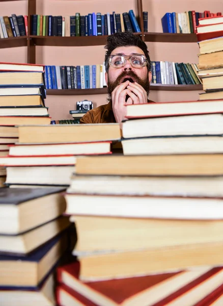 Mann mit nervösem Gesicht zwischen Bücherstapeln in der Bibliothek, Bücherregale im Hintergrund. Lehrer oder Schüler mit Bart tragen Brillen, sitzen mit Büchern am Tisch und sind defokussiert. wissenschaftliches Entdeckungskonzept — Stockfoto