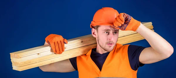 Construction worker carrying a wood plank on his shoulder. Carpenter wearing hard hat, protective gloves and safety orange vest, blue background. Protective equipment concept