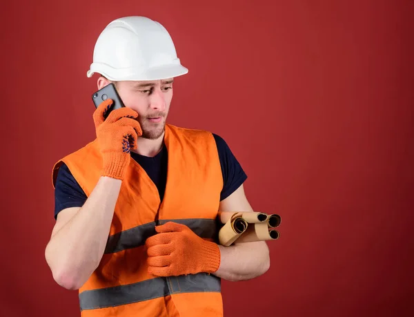 Engineer, architect, builder on busy face speaks on smartphone while holds blueprints. Man, foreman in helmet supervises construction on phone, red background. Architect concept