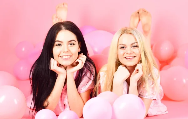 Las niñas yacían sobre el vientre cerca de globos, fondo rosa. Concepto de chismes. Rubia y morena en caras sonrientes se divierten en la fiesta del dormitorio doméstico. Hermanas o mejores amigos en pijama en fiesta de pijamas de niña — Foto de Stock