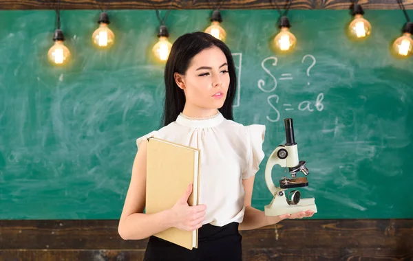 Dama en ropa formal con la cara tranquila en el aula. Concepto de biología. Profesor de biología tiene libro y microscopio. Lady científica sostiene libro y microscopio, pizarra en el fondo, espacio de copia — Foto de Stock