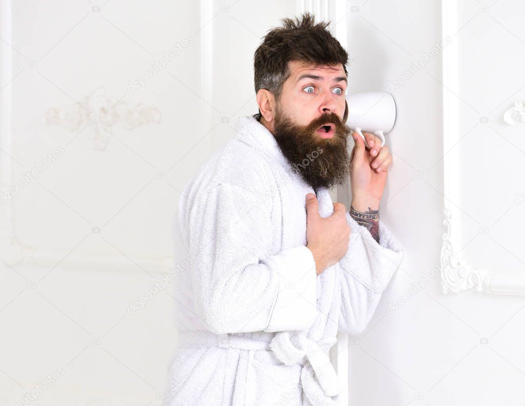 Man with beard and mustache eavesdrops using cup near wall. Hipster in bathrobe on surprised face secretly listen conversation. Privacy concept.