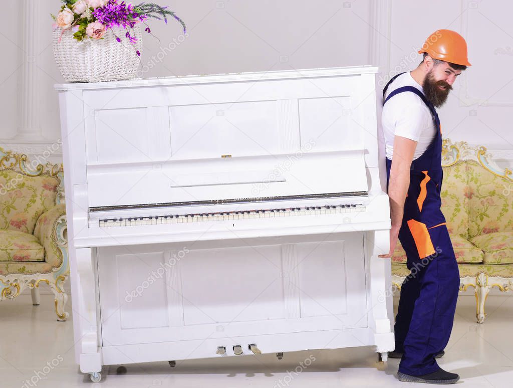 Loader moves piano instrument. Courier delivers furniture in case of move out, relocation. Man with beard, worker in overalls and helmet lifts up piano, white background. Delivery service concept
