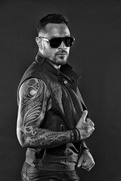 Tattooed man with biceps and triceps. Tattoo model with beard on unshaven face. Bearded man with tattoo on strong arms. Fashion macho in trendy sunglasses. Fashion style and trend, black and white