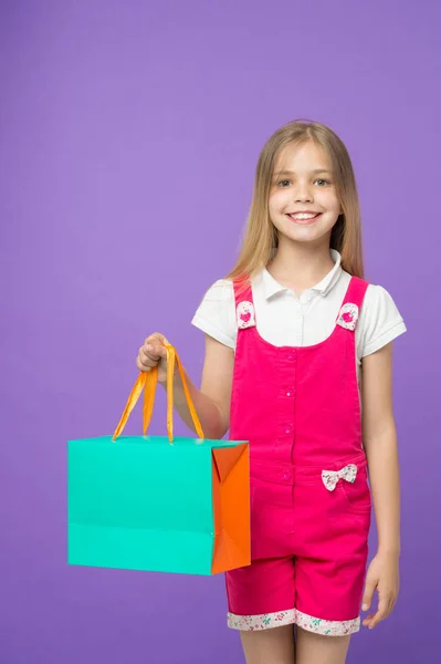 Kindje met boodschappentas op violette achtergrond. Gelukkig meisje glimlach met papieren zak. Kid shopper in mode jumpsuit. Feestdagen en viering. Tevreden met haar winkelen. verkoop en zwarte vrijdag — Stockfoto
