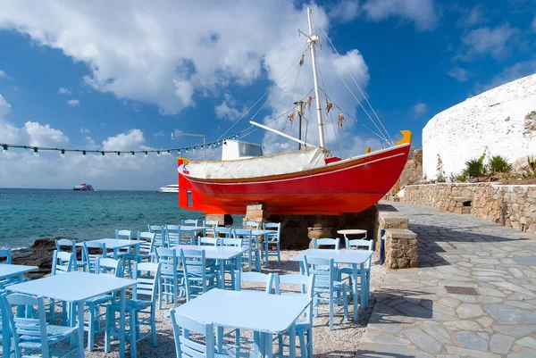 Ship and tavern furniture on quay in Mykonos, Greece. Red boat and blue tables on sea beach. Beach restaurant with sea view. Summer vacation on mediterranean island. Travelling for romance