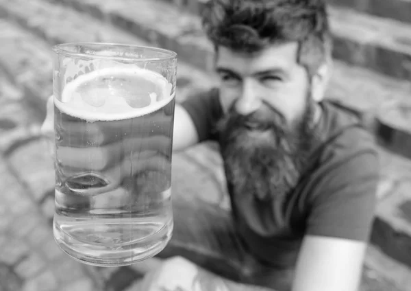 Um conceito de saúde. Homem com barba e bigode segura vidro com cerveja enquanto se senta em escadas de pedra, desfocado. Um tipo a levantar copos com cerveja. Hipster no rosto sorridente bebe cerveja ao ar livre — Fotografia de Stock