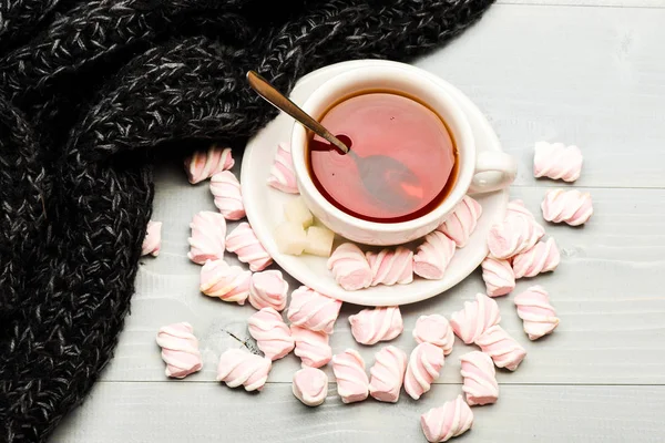 Cup with hot black tea and marshmallows. Coziness concept. Mug and sweets near cozy knitted scarf, top view. Mug filled with black brewed tea, spoon and heap of marshmallow on white wooden background — Stock Photo, Image