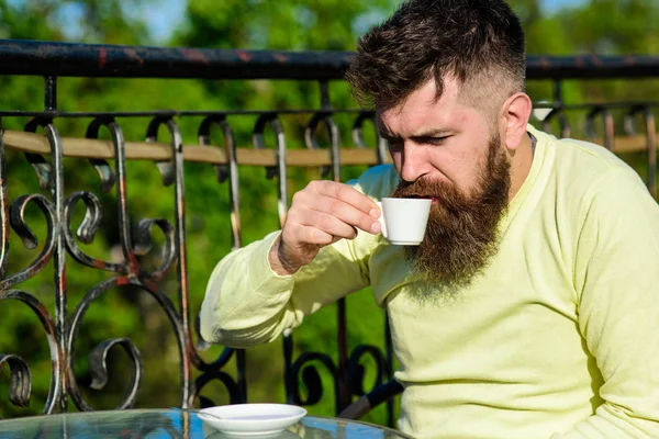 Bearded man with espresso cup, drinks coffee. Man with beard and mustache on strict face, fence of terrace on background, defocused. Man with long beard looks strict and serious. Coffee break concept.