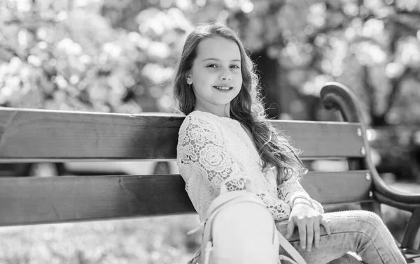 Menina no rosto sorridente senta-se no banco, árvore sakura no fundo, desfocado. Criança bonito com cabelo longo e bonito desfrutar de dia de primavera ensolarado. Menina relaxante no parque perto de flor de cereja. Conceito de beleza natural — Fotografia de Stock