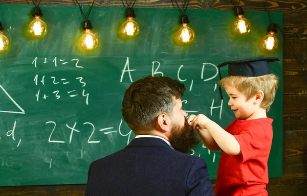 Individual education concept. Little boy playing with beard of man next to him. Adult turned back and smiling child in graduation cap side view. Young guy and small kid in front of the green board — Stock Photo, Image