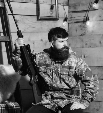Sakallı adam ahşap iç arka planda kamuflaj elbiseler giyer. Avcı, av için hazır elinde tabanca ile acımasız hipster. Hakem kavramı. Maço gamekeepers evinde sıkı yüzünde