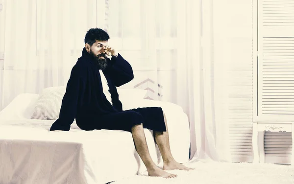 Un tipo con cara dormilona levantándose por la mañana. Macho con barba y bigote lento relajante después de la siesta, descanso. El hombre en bata se sienta en la cama, cortinas blancas en el fondo. Concepto de despertar — Foto de Stock