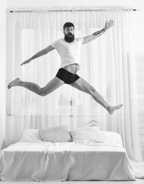 Man in shirt and underpants jumping on bed, white curtains on background. Macho with beard jumps high in air. Full of strength and energy concept. Guy on cheerful face full of energy in morning