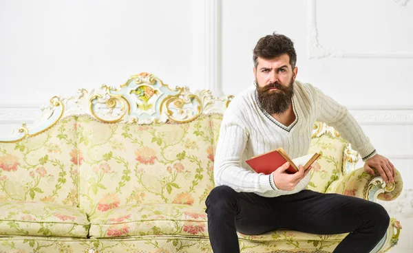 Hombre con barba y bigote se sienta en el sofá de estilo barroco, sostiene libro, fondo blanco de la pared. Reflexiones sobre el concepto de literatura. Un tipo pensando en literatura. Macho en estricta cara terminó libro de lectura —  Fotos de Stock