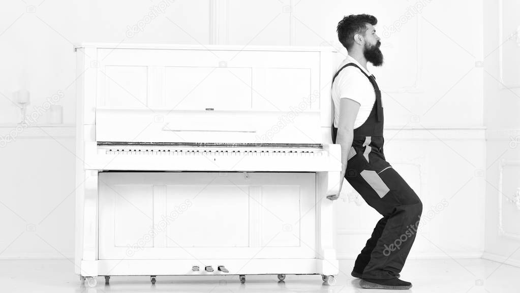 Loader moves piano instrument. Man with beard and mustache, worker in overalls lifts up piano, white background. Courier delivers furniture in case of move out, relocation. Delivery service concept