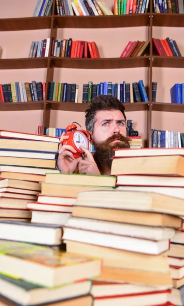 Profesor o estudiante con barba estudiando en la biblioteca. Hombre, científico asomándose de montones de libros con despertador. Un hombre atento a la cara escuchando el reloj, estanterías en el fondo. Concepto de parada temporal —  Fotos de Stock