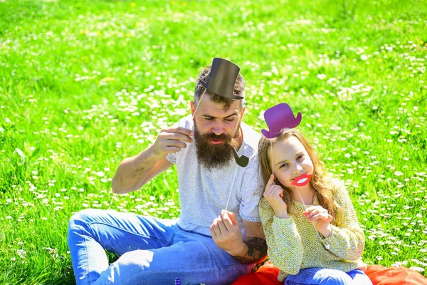 Aristocrates concept. Family spend leisure outdoors. Dad and daughter sits on grass at grassplot, green background. Child and father posing with top hat, lips and pipe photo booth attributes — Stock Photo, Image