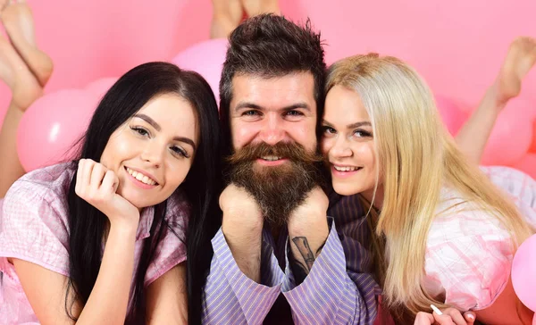 Girls fall in love with bearded macho, pink background. Threesome on smiling faces lay near balloons. Alpha male concept. Man with beard and mustache attracts blonde and brunette girls