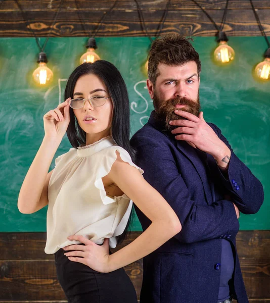 Sakal ve gözlük stand sırt sırta, kara tahta arka plan öğretmen olan adam. Öğretmen ve öğretmen rahat görünüyor. Ekip çalışması kavramı. Bayan ve birlikte okulda çalışan hipster