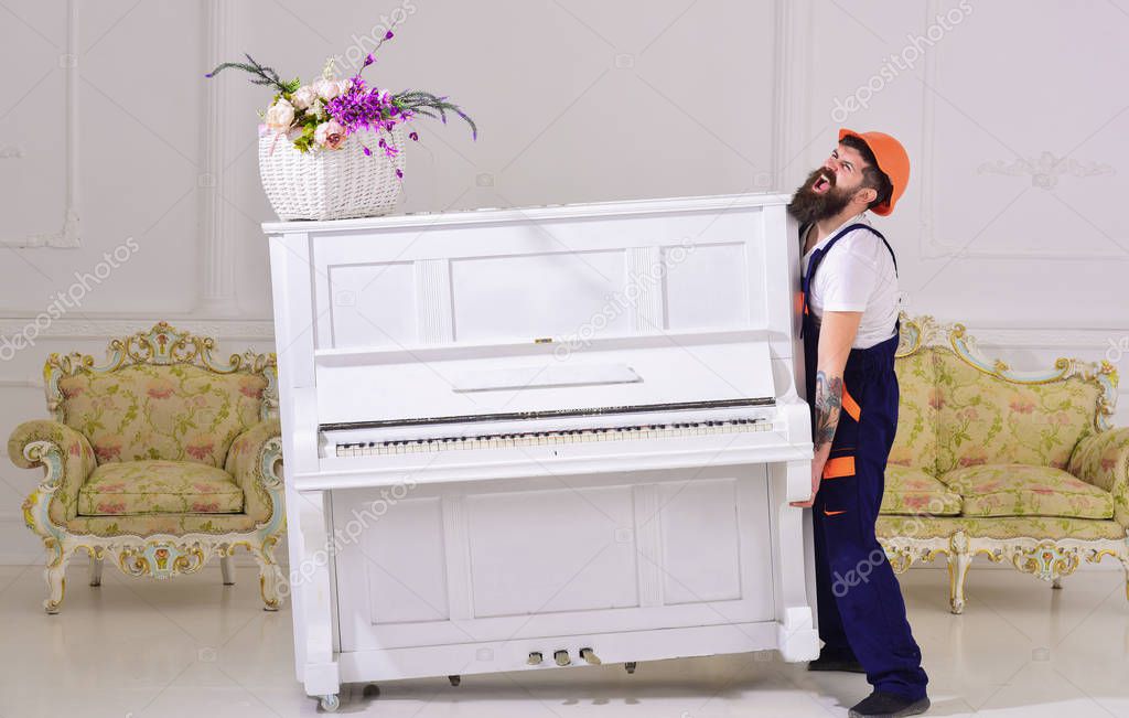 Courier delivers furniture, move out, relocation. Heavy loads concept. Man with beard worker in helmet and overalls lifts up, efforts to move piano, white background. Loader moves piano instrument