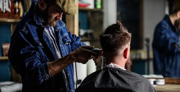 Barber with clipper trimming hair of client, rear view. Hipster client getting haircut. Hipster lifestyle concept. Barber with hair clipper works on haircut of bearded guy, barbershop background — Stock Photo, Image