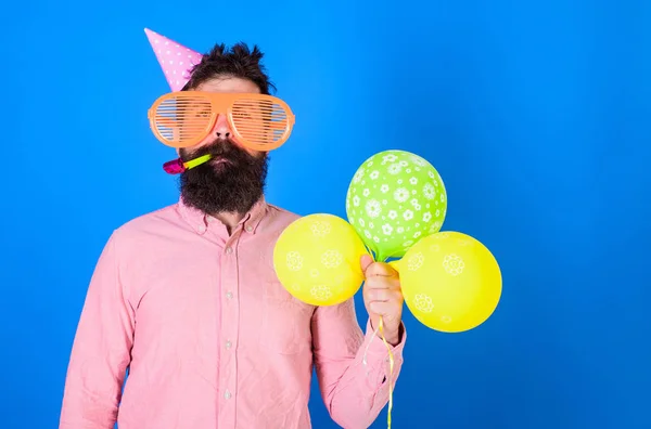 Conceito de celebração. Hipster em óculos de sol gigantes comemorando aniversário. Homem com barba e bigode no rosto calmo sopra em chifre de festa, fundo azul. Cara em chapéu de festa com balões de ar celebra — Fotografia de Stock