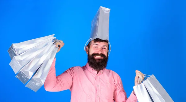 Mann mit modischem Bart und lächelndem Gesicht isoliert auf blauem Hintergrund. Hipster im rosafarbenen Hemd beim Einkaufen, großer Spender. glücklicher bärtiger Mann, der silberne Taschen trägt und eine als Hut trägt, Einkaufskonzept — Stockfoto