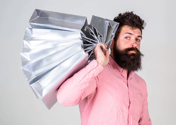 Concepto de compras. Chico de compras en temporada de ventas, lleva un montón de bolsas en el hombro. Hombre con barba y bigote sostiene bolsas de compras, fondo gris. Hipster en serio la cara de compras —  Fotos de Stock