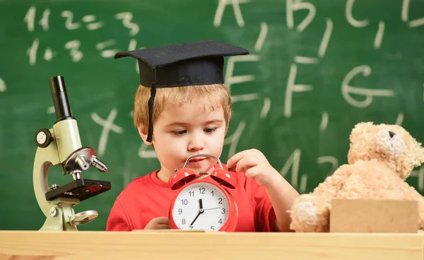 Kid boy i akademiska cap nära mikroskopet, rymmer klockan i klassrummet, svarta tavlan på bakgrund. Elev väntar skolan paus. Barn på sorgsna ansikte ser väckarklocka. Skolan paus koncept — Stockfoto