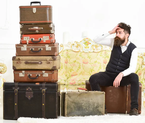 Hombre con barba y bigote lleno de equipaje, fondo interior blanco. Macho elegante en la cara cansada se sienta, agotado al final del embalaje, cerca de la pila de maletas vintage. Concepto de equipaje y reubicación —  Fotos de Stock