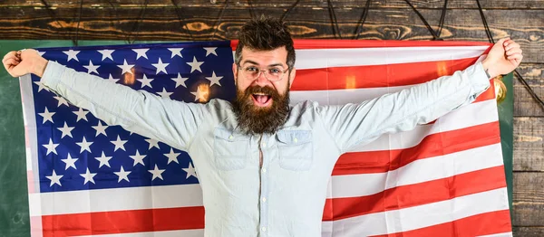 American educational system concept. Student exchange program. Man with beard and mustache on happy face holds flag of USA, wooden background. American teacher in eyeglasses holds american flag