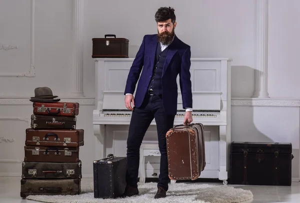 Baggage delivery concept. Macho elegant on strict face stands near pile of vintage suitcase, holds suitcase. Man, traveller with beard and mustache with luggage, luxury white interior background