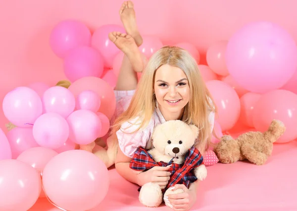 Woman cute celebrate birthday with balloons. Girl in pajama, domestic clothes lay near air balloons, pink background. Birthday girl concept. Blonde on smiling face relaxing with teddy bear toy