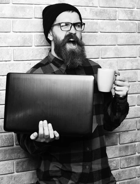 Bearded brutal caucasian hipster holding laptop with cup — Stock Photo, Image