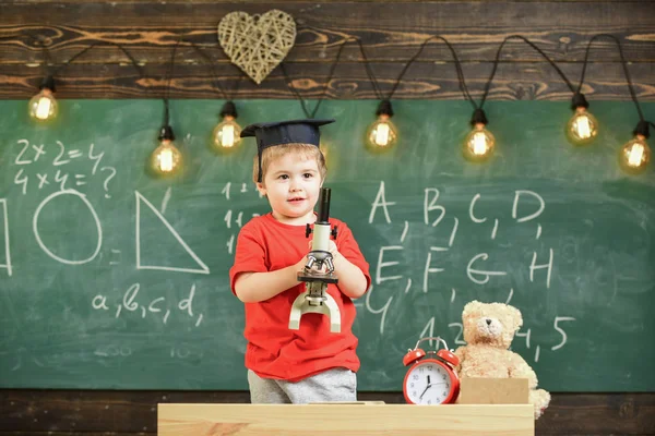 Smart kid concept. First former interested in studying, learning, education. Child on happy face holds microscope. Kid boy in academic cap work with microscope in classroom, chalkboard on backgroun — Stock Photo, Image