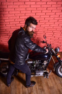 Yenilikçi, vahşi Motorcu deri ceket ciddi yüzüne üzerinde motosiklet üzerinde oturur. Erkeksi hobi kavramı. Sakallı, motorcu motorlu bisiklet garaj, tuğla duvar arka plan yakınındaki deri ceketli adam