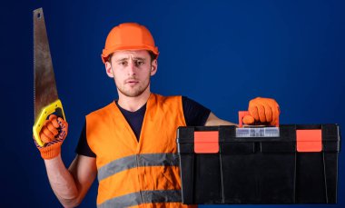 Marangoz kavramı. Kask, kask içinde adam araç kutusu taşır ve el testeresi, mavi arka plan tutar. İşçi, repairer, tamirci üzerinde ciddi bir yüz taşıyan araç, onarım için kopya alanı hazır