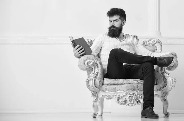 Hombre con barba y bigote se sienta en el sillón y la lectura, fondo blanco de la pared. Macho pasa el ocio con el libro. Conocedor, en la cara seria disfrutar de la literatura. Conocedor del concepto de literatura —  Fotos de Stock