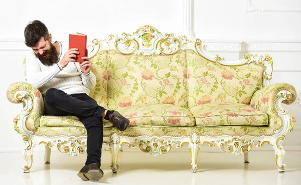 A fickó élvezet régi könyvet olvasott. Humoros irodalom fogalma. Férfi szakáll és bajusz ül a barokk stílusú kanapé, tartja a könyvet, fehér háttér. Macho-a nevető arc könyvet olvas — Stock Fotó