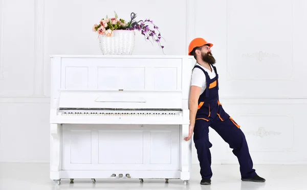 Hombre con barba trabajador en casco y overoles empuja, esfuerzos para mover el piano, fondo blanco. El mensajero entrega muebles, se muda, se traslada. El cargador mueve el instrumento de piano. concepto de cargas pesadas — Foto de Stock