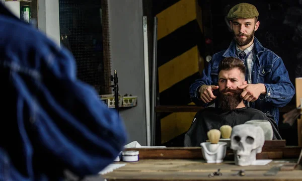 Cliente Hipster com corte de cabelo fresco ou penteado. Reflexão de bigode barbeiro styling de cliente com cera por mãos. Homem com barba e bigode na cadeira de cabeleireiro, espelho de fundo. Conceito de barbeiro — Fotografia de Stock