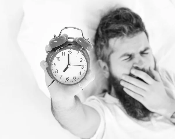 El hombre con la cara bostezante yace en la almohada, sostiene el despertador . —  Fotos de Stock