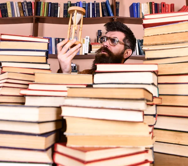 O homem no rosto pensativo mantém a ampulheta enquanto estuda, estantes de livros no fundo. Professor ou estudante com barba estudando na biblioteca. Conceito de fluxo de tempo. Homem, cientista de óculos olha para ampulheta — Fotografia de Stock