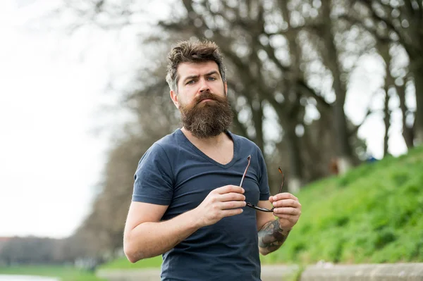 Killen ser cool med snygga solglasögon. Ögon skydd koncept. Mannen med skägg och mustasch innehar solglasögon, Oskarp bakgrund. Hipster på säker ansikte med moderiktiga solglasögon — Stockfoto